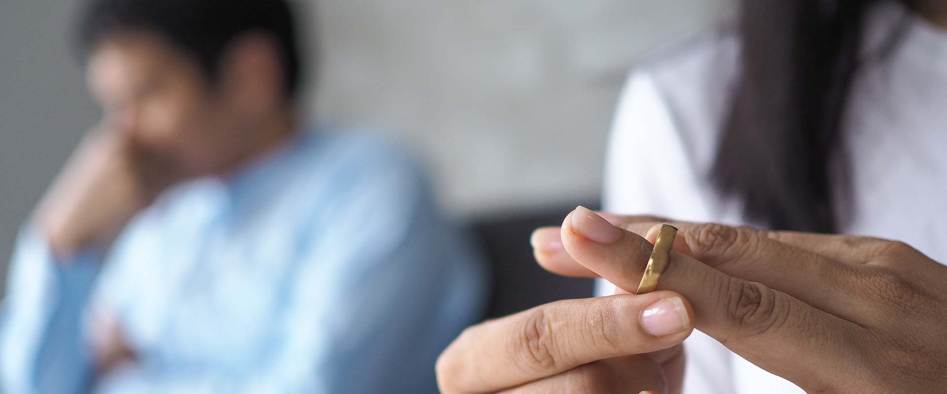 Anerkennung ausländischer Ehescheidungen in Deutschland, eine Frau nimmt den Ehering vom Finger, im Hintergrund sitzt ihr verzweifelter Mann